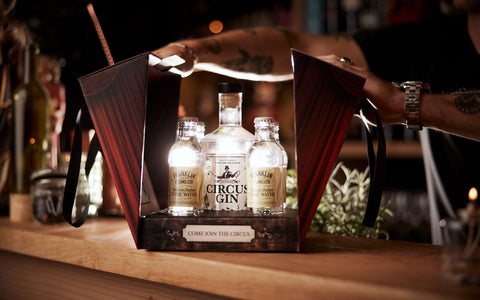 En person åbner en gaveæske med en flaske Circus Gin og to tonicflasker på en bardisk af træ, hvor der står COME JOIN THE CIRCUS på etiketten.