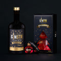 En flaske GINSTR Winter Gin fra Stuttgart Distillery står ved siden af en "GINSTR Winter Gin & Dry Gin Julekugler"-gaveæske med tre røde, ginfyldte ornamenter og et sort armbånd, alt sammen på en sort baggrund.