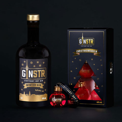 En flaske GINSTR Winter Gin fra Stuttgart Distillery står ved siden af en "GINSTR Winter Gin & Dry Gin Julekugler"-gaveæske med tre røde, ginfyldte ornamenter og et sort armbånd, alt sammen på en sort baggrund.
