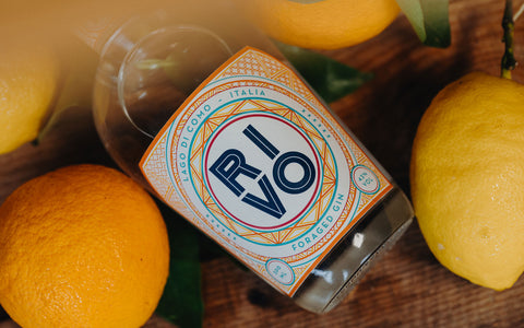 En flaske RIVO-gin ligger på en træplade omgivet af friske appelsiner og citroner. På etiketten står der "RIVO Lago di Como Italia Foraged Gin" med fede bogstaver og farverige geometriske mønstre.