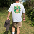 En person går på en græsklædt sti udendørs iført en T-shirt med teksten »Magi Spirits RIVO Gin – Keep Discovering« og et farverigt motiv, beige shorts, hvide sokker, sneakers og en sort kasket, mens vedkommende holder en mørk sweatshirt i hånden. Stien er omgivet af træer og buske.