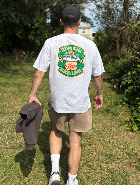 En person går på en græsklædt sti udendørs iført en T-shirt med teksten »Magi Spirits RIVO Gin – Keep Discovering« og et farverigt motiv, beige shorts, hvide sokker, sneakers og en sort kasket, mens vedkommende holder en mørk sweatshirt i hånden. Stien er omgivet af træer og buske.