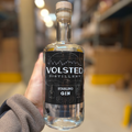 En hånd holder en flaske Volsted Starling Gin fra Volsted Distillery i et lagerlokale, hvor hylder og kasser er let slørede i baggrunden.