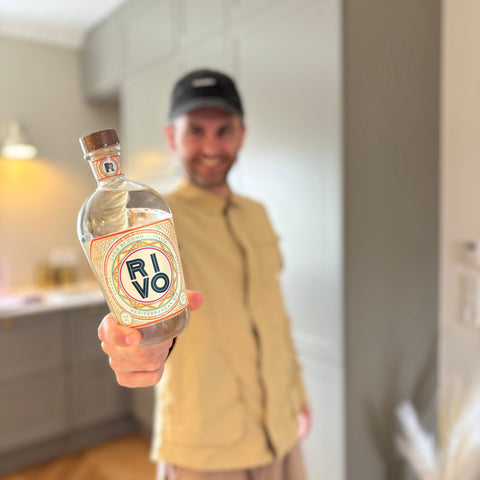 En person iført en beige skjorte og en sort kasket holder en flaske RIVO Mediterranean Gin fra Magi Spirits frem mod kameraet, mens vedkommende står i et moderne køkken med skabe i neutrale farver i baggrunden. Scenen fremhæver denne gins friske karakter, der er beriget med italienske citrusfrugter – perfekt til at nyde de forfriskende øjeblikke.
