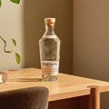 En klar glasflaske Stauning Danish Dry Gin by Stauning Whisky med træprop står på et lyst træbord ved siden af en grøn plante i et varmt lys - en indbydende ramme for at nyde denne fine danske gin.
