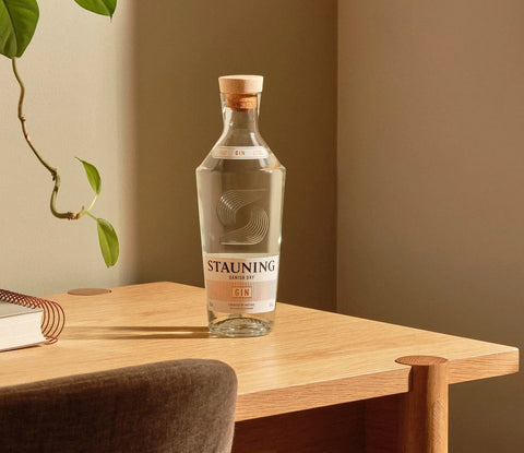 En klar glasflaske Stauning Danish Dry Gin by Stauning Whisky med træprop står på et lyst træbord ved siden af en grøn plante i et varmt lys - en indbydende ramme for at nyde denne fine danske gin.