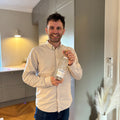 En smilende mand står i et moderne køkken med en gennemsigtig flaske Stalinskaya-vodka i hånden, iført en lys skjorte og jeans. I forgrunden, ved siden af en dekorativ vase med tørret græs, står en flaske Stauning Danish Dry Gin fra Stauning Whisky, der tilfører det moderne miljø et håndværksmæssigt præg.