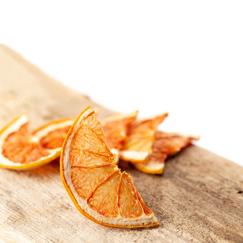 Flere tørrede grapefrugter fra Frugtskiver.dk er arrangeret på en rustik træoverflade mod en hvid baggrund med fokus på den forreste skive, der fremhæver frugtens strukturerede, rynkede overflade.