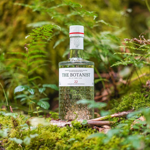 En flaske The Botanist Islay Dry Gin står på grønt mos og er omgivet af bregner i en frodig skov.