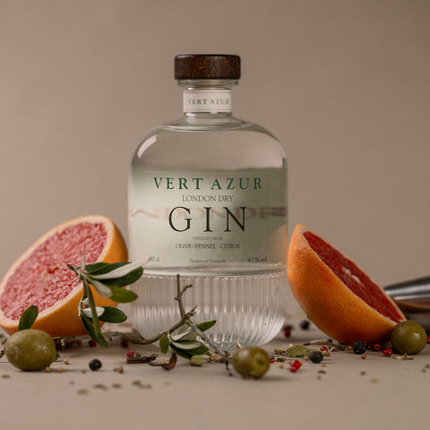 En flaske Vert Azur Gin fra Vert Azur er placeret på en neutral baggrund, omgivet af halve grapefrugter, grønne oliven, olivengrene og spredte peberkorn.