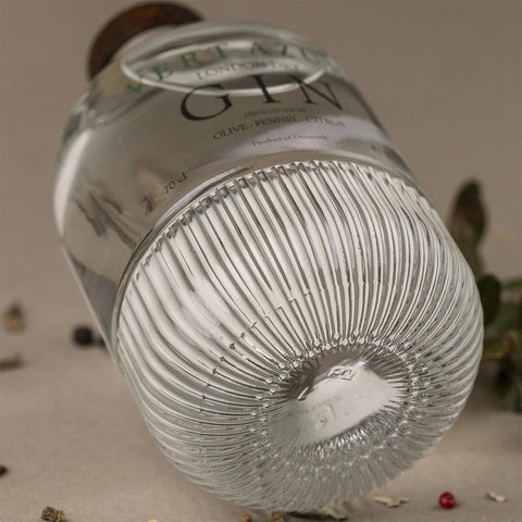 Et nærbillede af en tekstureret Vert Azur Gin-flaske fra Vert Azur, der ligger på siden med "GIN" på etiketten. Krydderurter og peberkorn er spredt i nærheden mod en blødt sløret, neutral baggrund.